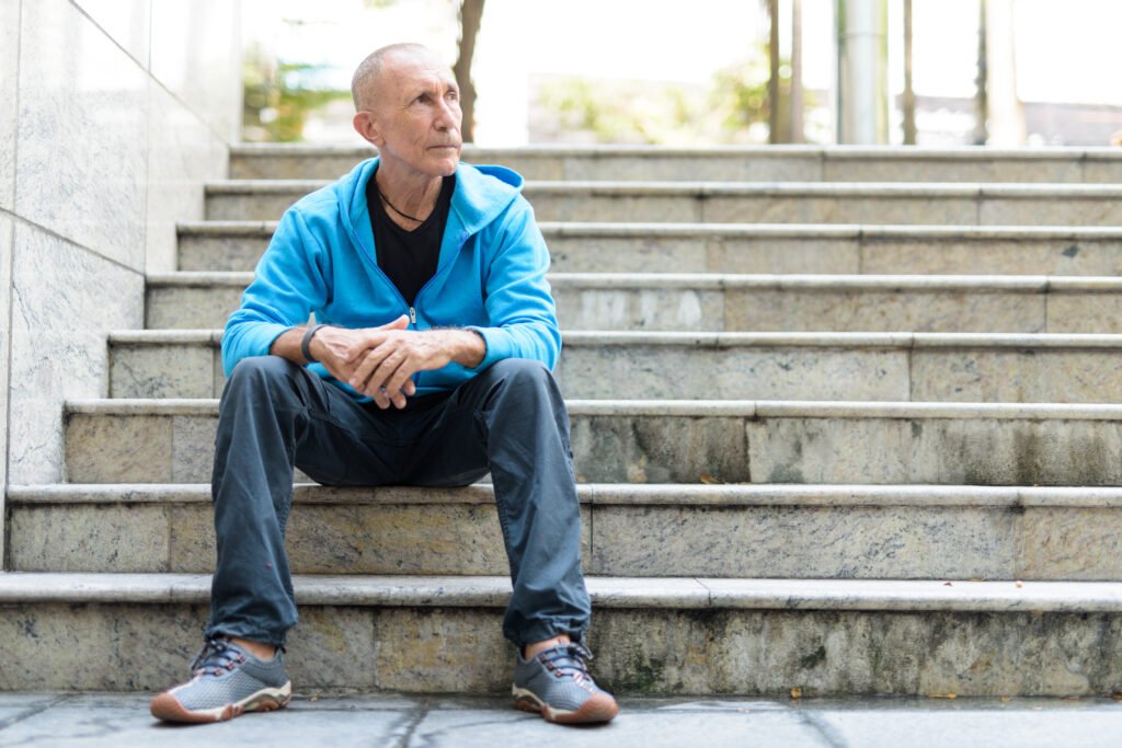 A senior man with a shaved head, wearing a bright blue zip-up hoodie, black t-shirt, and dark jeans, sits on stone steps outdoors, looking off to the right with a contemplative expression.