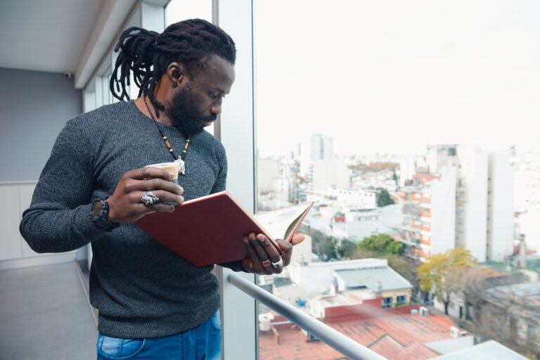 Black writer with dreadlocks reading a notebook and drinking coffee while overlooking a cityscape