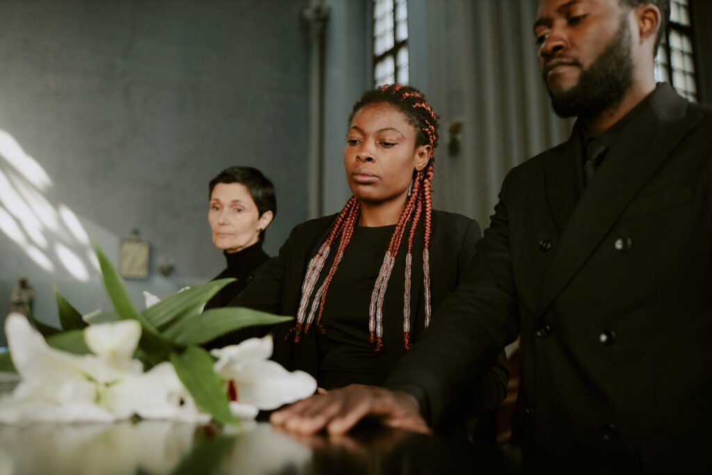 Mourners standing with somber expressions at a funeral service, with white lilies visible in the foreground